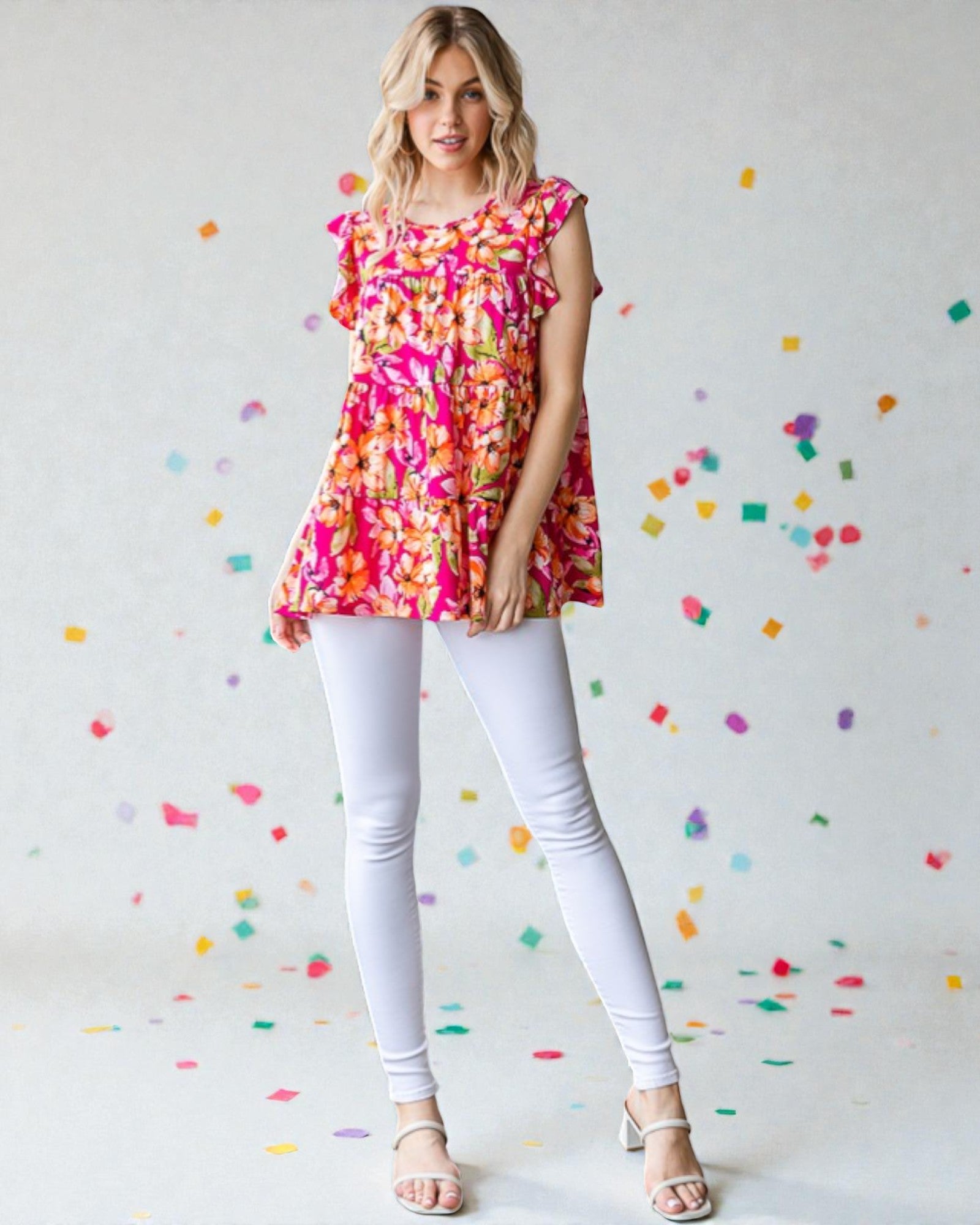 Woman wearing a colorful floral top and white pants against a confetti-patterned wall.