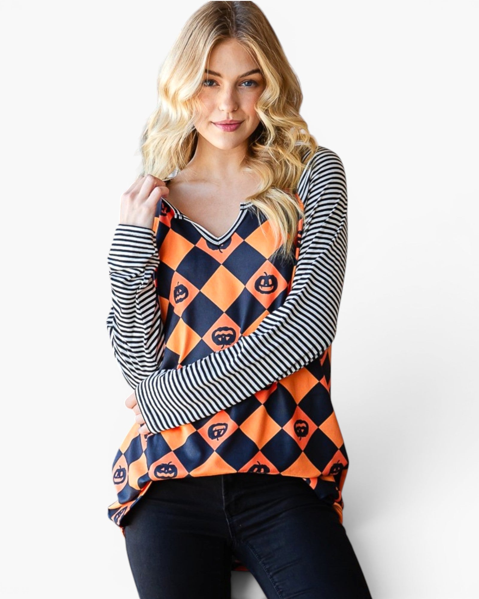 A woman modeling a long-sleeve top with a navy and orange harlequin pattern featuring small jack-o'-lantern motifs, layered over a black and white striped long-sleeve shirt, paired with black jeans against a plain white background.