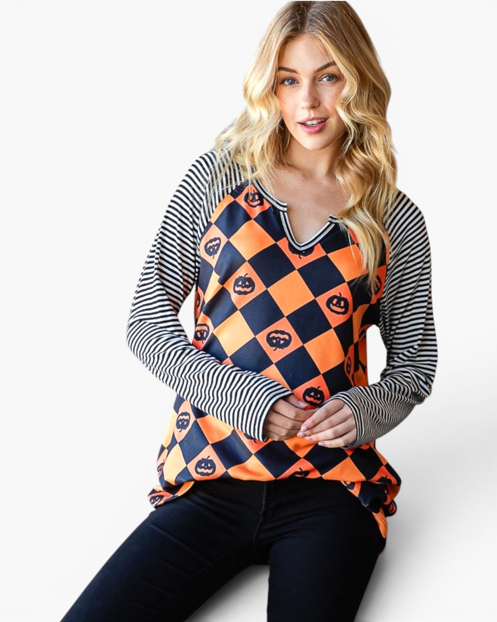 A woman wearing a long-sleeve Halloween top with a navy and orange harlequin pattern featuring jack-o'-lantern motifs, paired with black pants, against a light gray studio background.