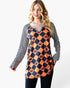 A woman modeling a long-sleeve tunic top with a Halloween theme. The top features a black and orange harlequin diamond pattern with small jack-o'-lantern faces, combined with black and white striped sleeves.