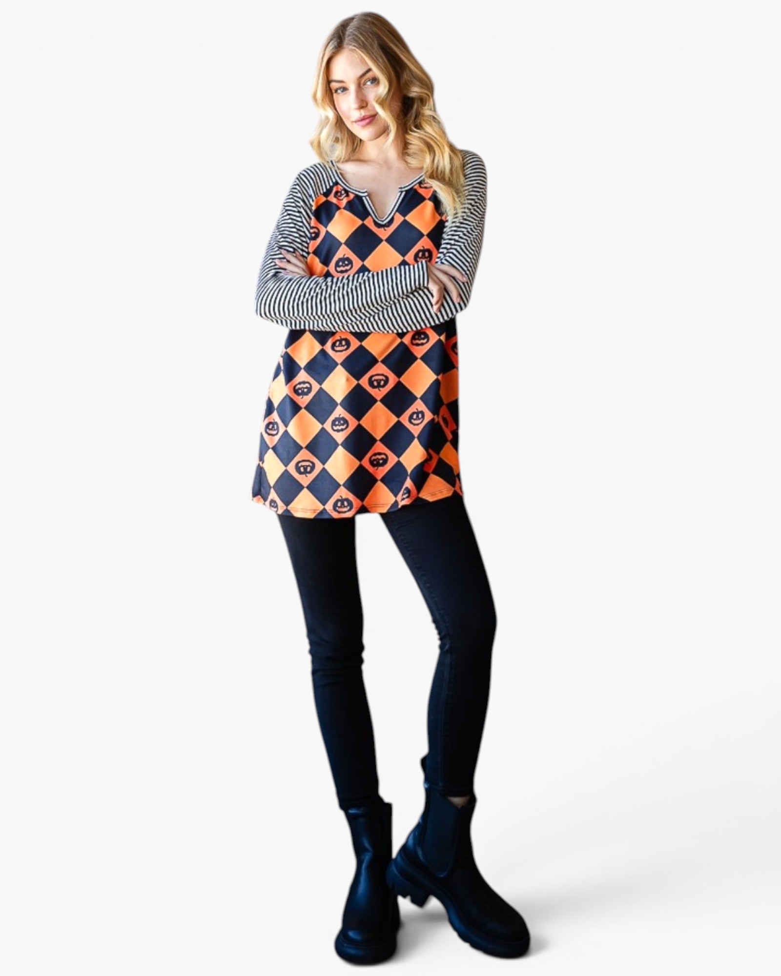 A woman modeling a long-sleeve tunic top with a navy and orange harlequin diamond pattern over a black and white striped long-sleeve shirt, paired with black leggings and dark blue ankle boots, against a light gray background.