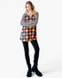 A woman modeling a long-sleeve tunic top with a navy and orange harlequin diamond pattern over a black and white striped long-sleeve shirt, paired with black leggings and dark blue ankle boots, against a light gray background.