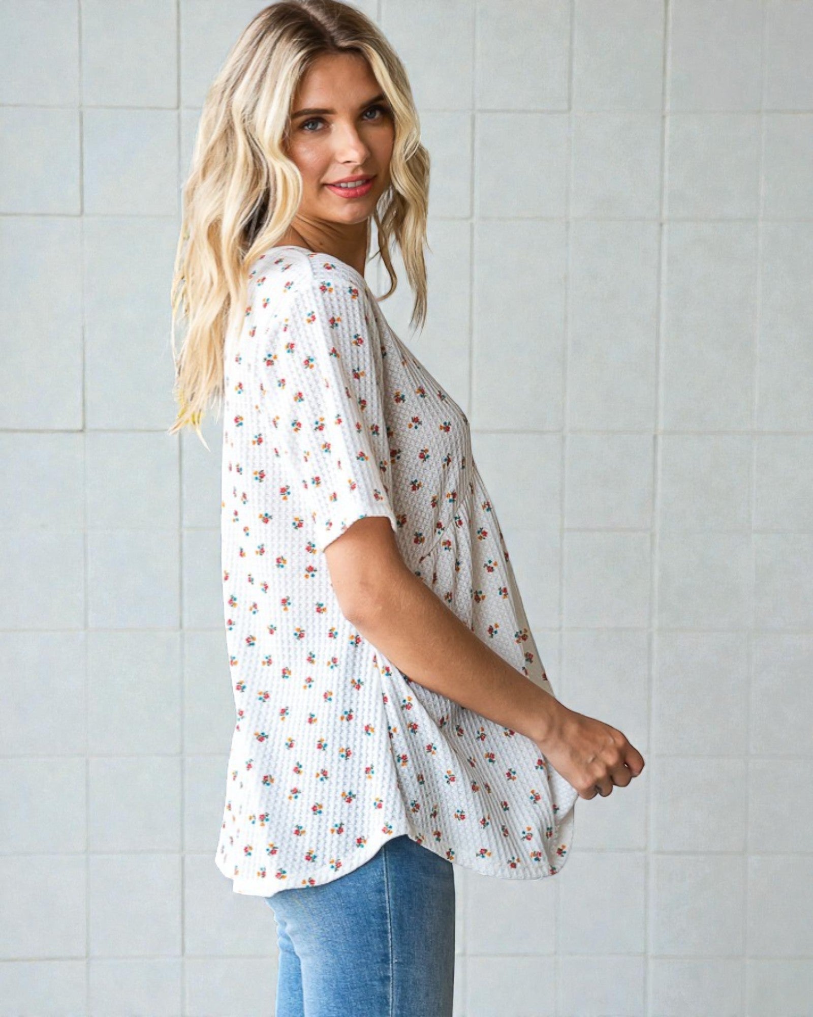 Woman wearing a white floral blouse and blue jeans against a light tiled wall.