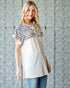 Woman wearing a white top with a patterned sleeve against a textured wall.