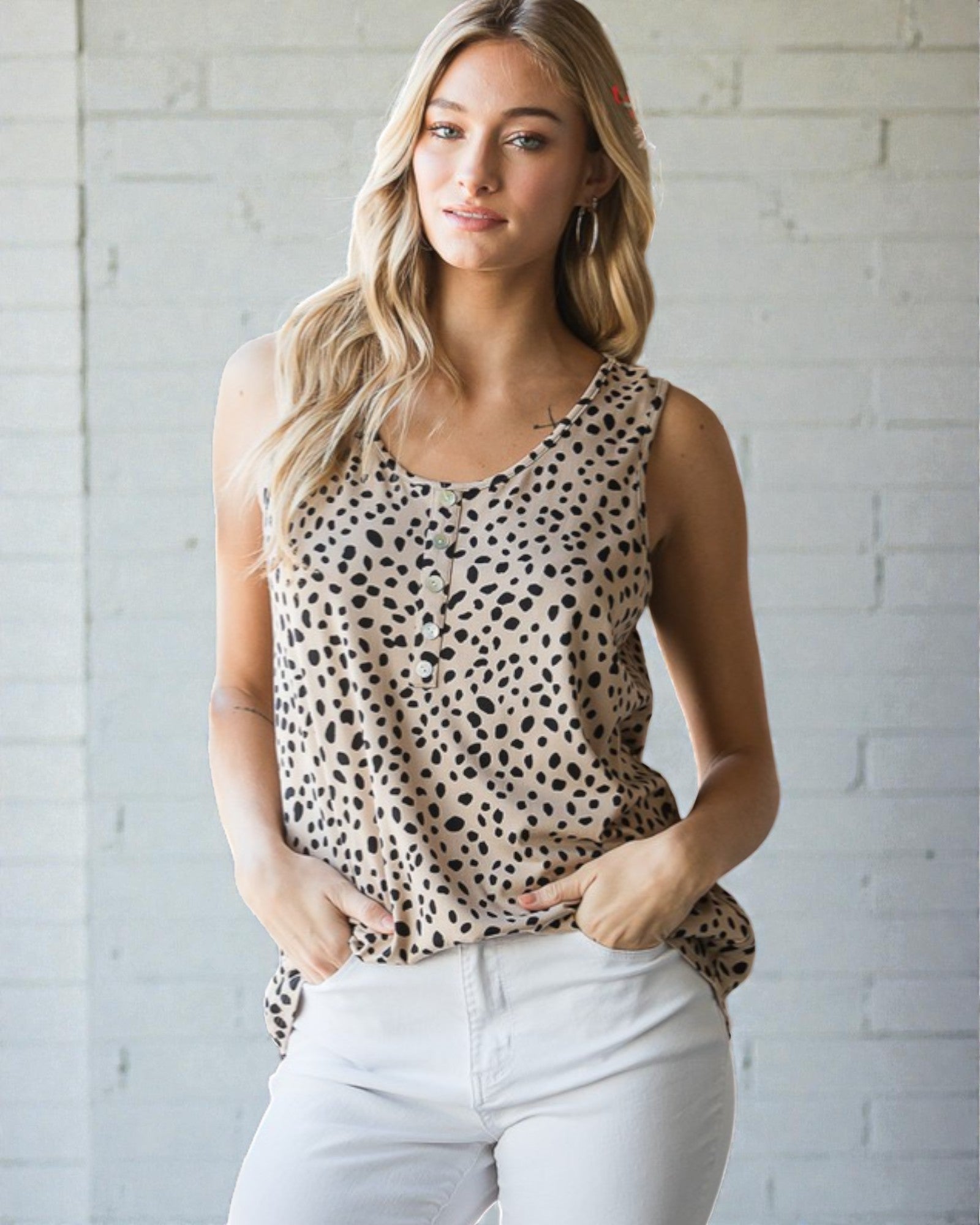 Woman wearing a leopard print tank top and white pants against a light brick wall.