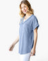 A woman wearing a light blue and white vertically striped short-sleeve V-neck top, paired with white pants, photographed against a solid white studio background.