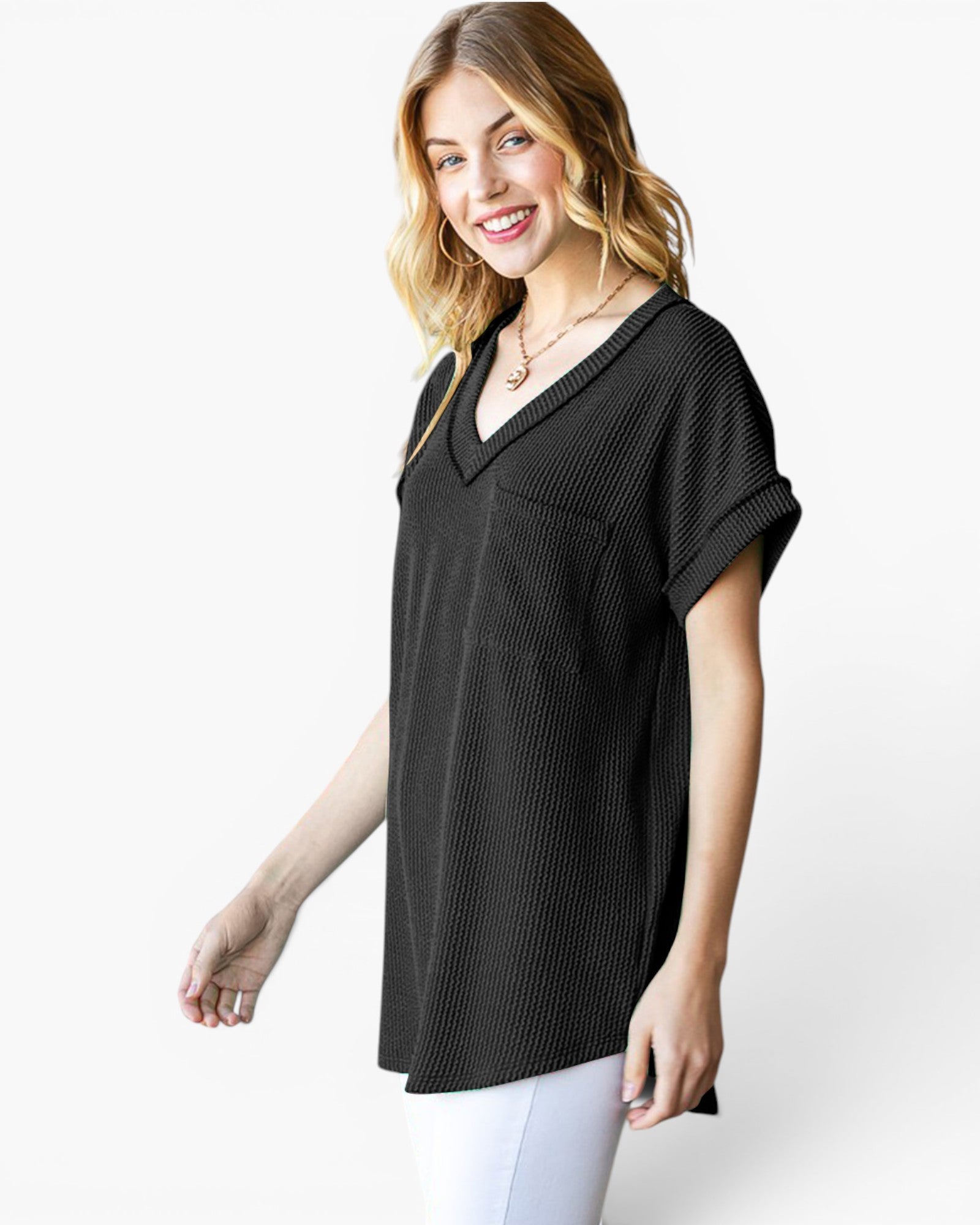 A woman wearing a black short-sleeve V-neck top with a textured vertical rib pattern and a chest pocket, paired with white pants, against a light gray studio background.