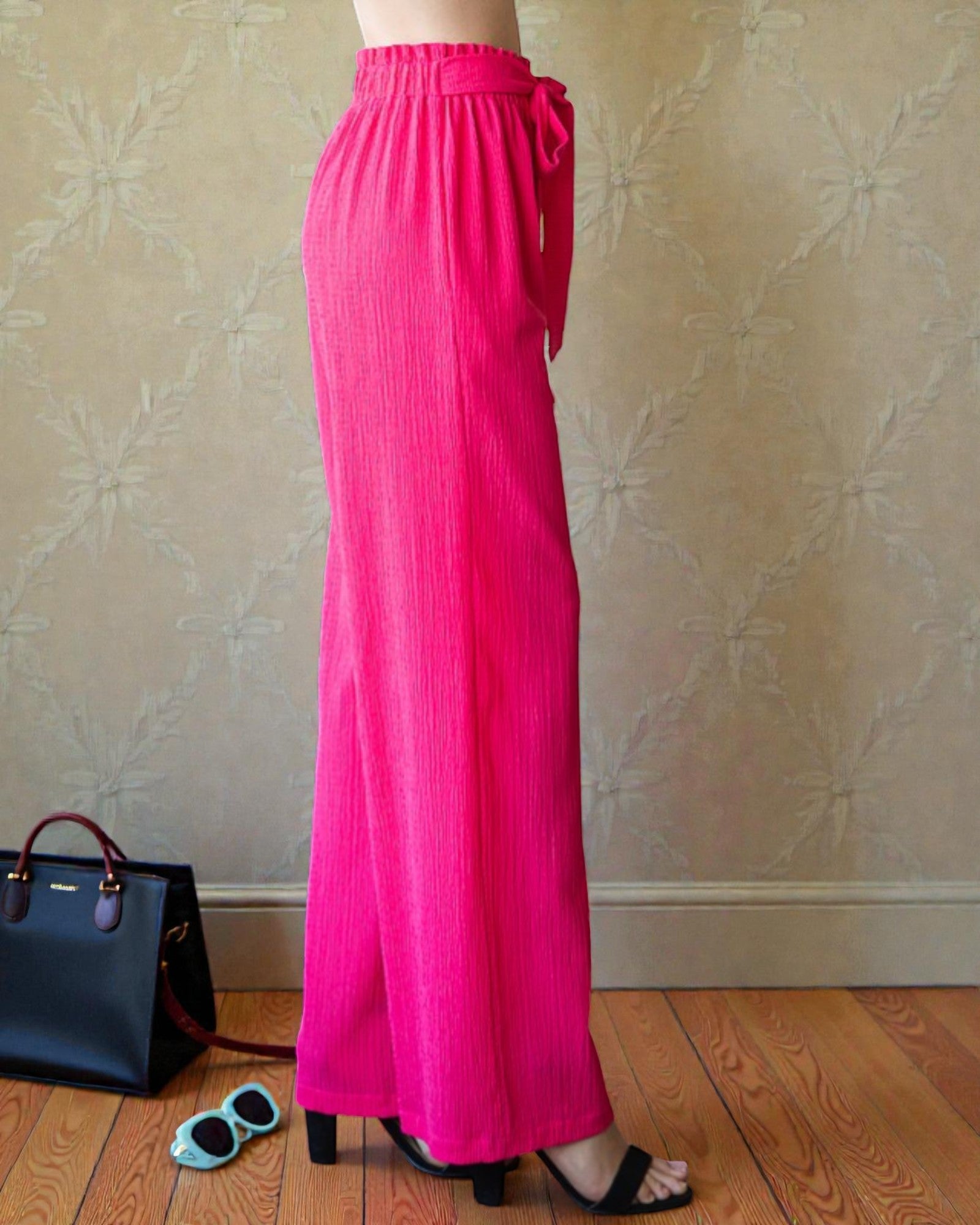 Person wearing bright pink wide-leg pants standing against a patterned wall with a handbag and sunglasses on the floor.