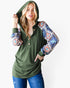 A woman wearing an olive green hooded henley top with patterned, colorful sleeves and blue jeans, posed against a plain white studio background.