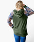 A woman shown from the back wearing an olive green hooded long-sleeve top with patterned raglan sleeves featuring a Southwestern geometric design in blue, red, and beige, paired with blue jeans against a white studio background.
