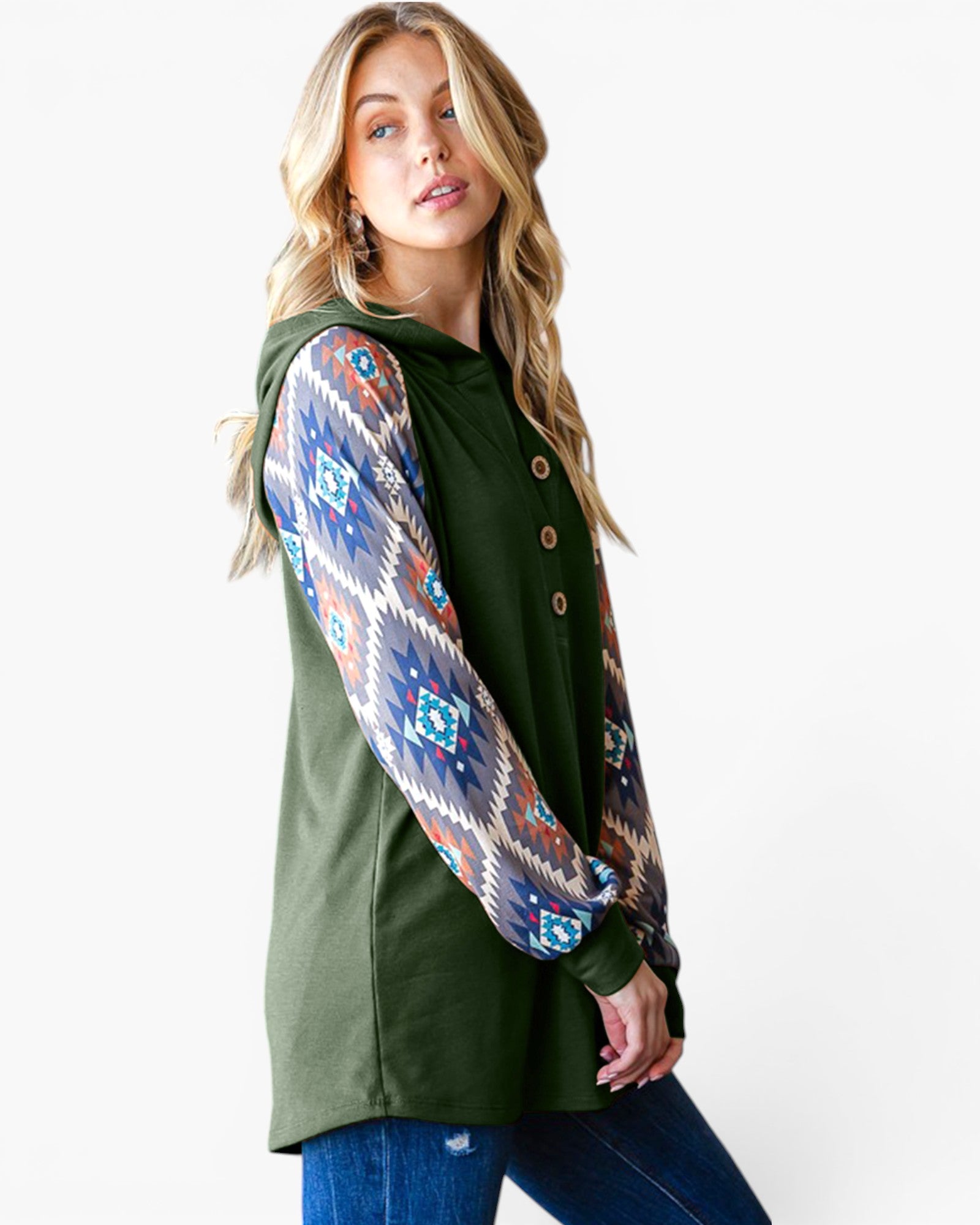 A woman wearing an olive green hooded tunic top with long patterned sleeves featuring a colorful Southwestern geometric print, paired with blue jeans, against a plain white studio background.
