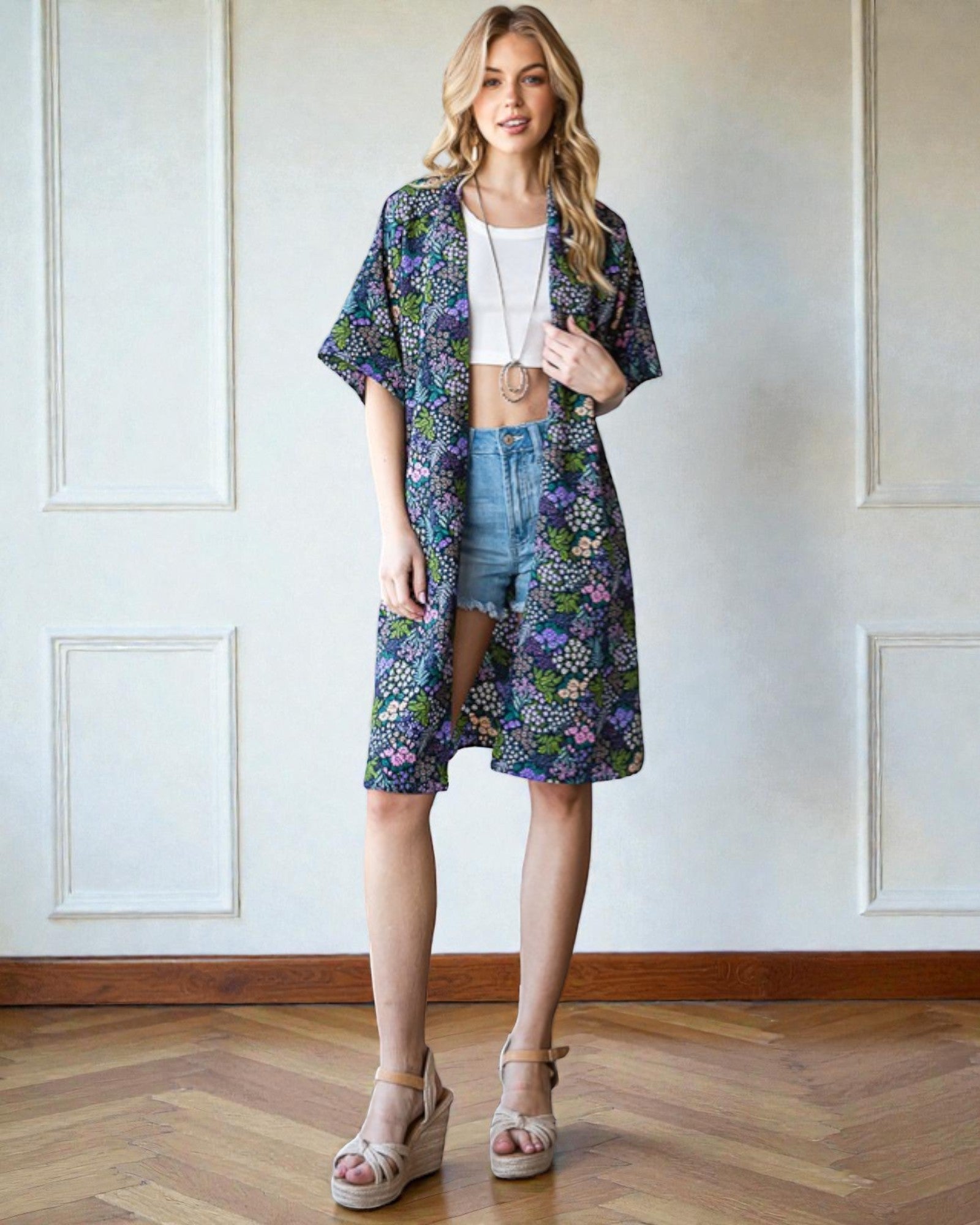 Woman wearing a floral kimono over a white top and denim shorts in a room with wooden flooring and white walls.