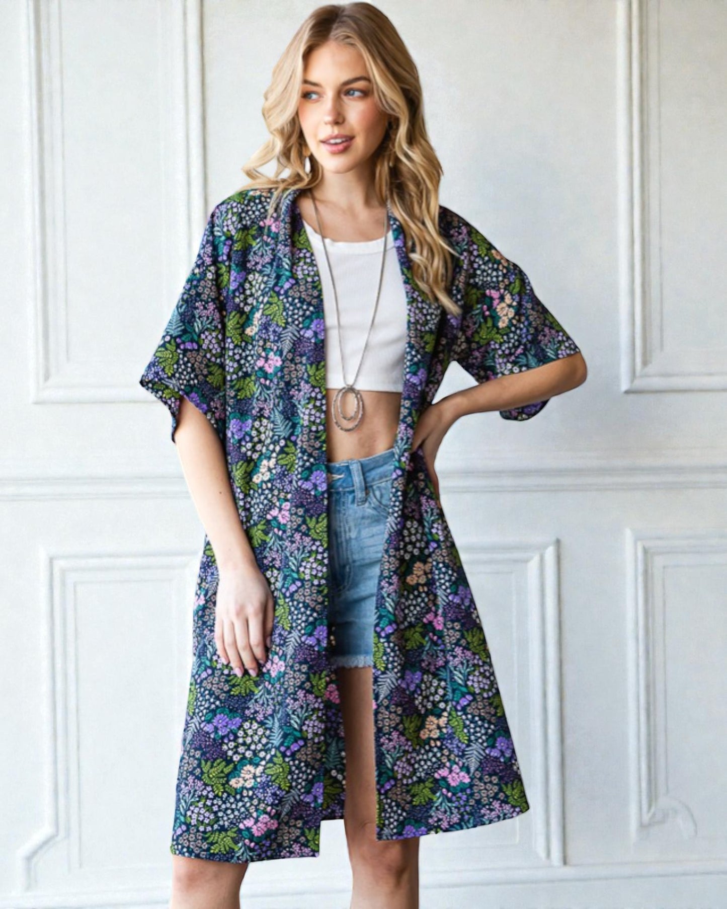 Woman wearing a floral kimono over a white top and denim shorts against a white wall.