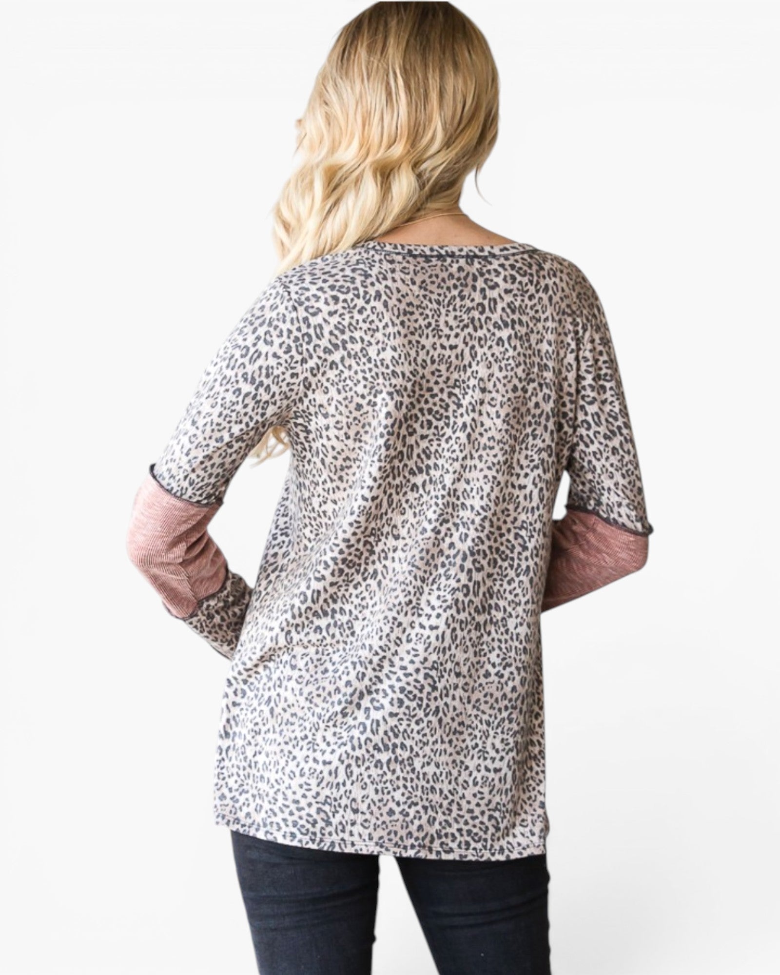 A woman viewed from the back wearing a leopard-print tunic top with dusty rose three-quarter sleeves and dark jeans, against a plain white background.