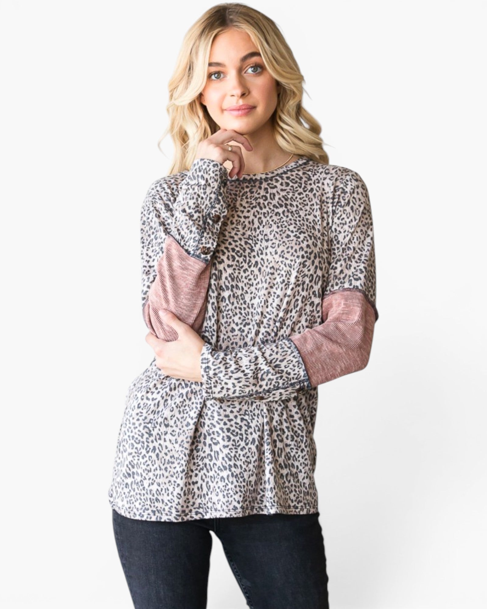 A woman modeling a long-sleeve top with a beige, brown, and black leopard print pattern and dusty rose pink elbow patches, paired with dark jeans against a plain white background.