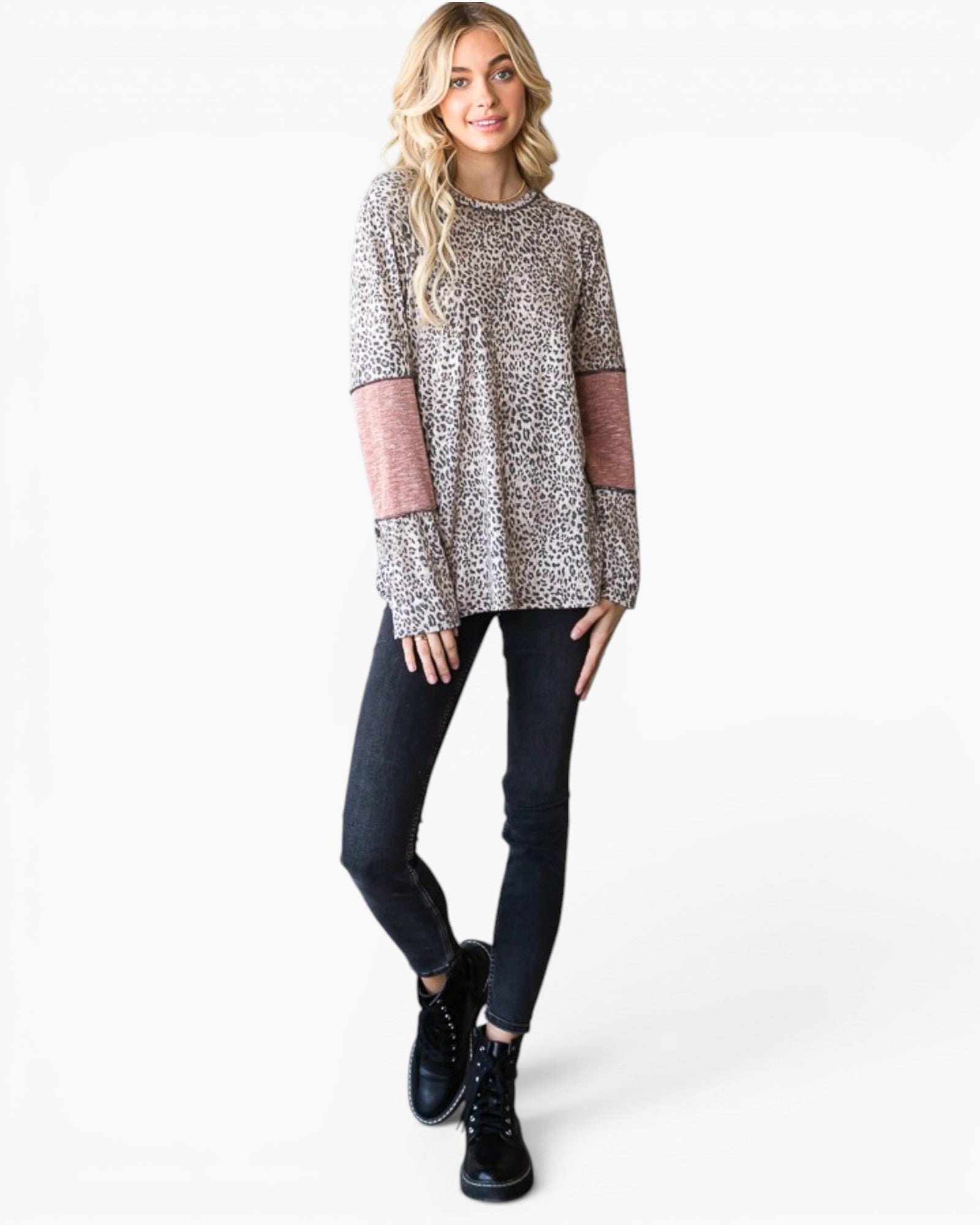 A woman modeling a long-sleeve knit top with a beige and black leopard print, dusty pink elbow patches, and flared cuffs, paired with black jeans and black lace-up boots against a white studio background.