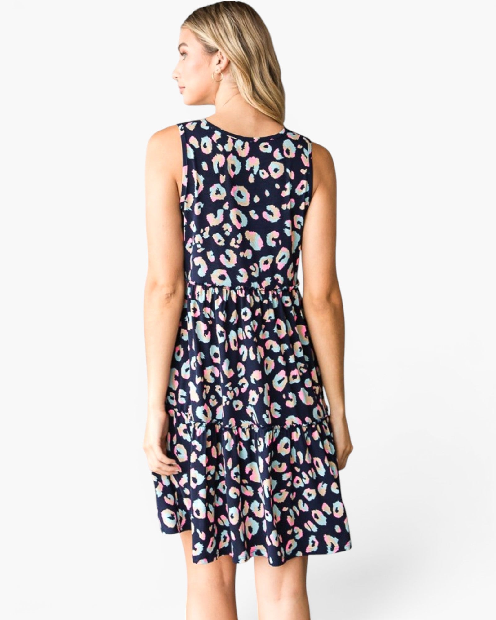 A woman modeling a sleeveless tiered dress from the back, featuring a navy blue base with a colorful pastel leopard print pattern, against a plain white studio background.
