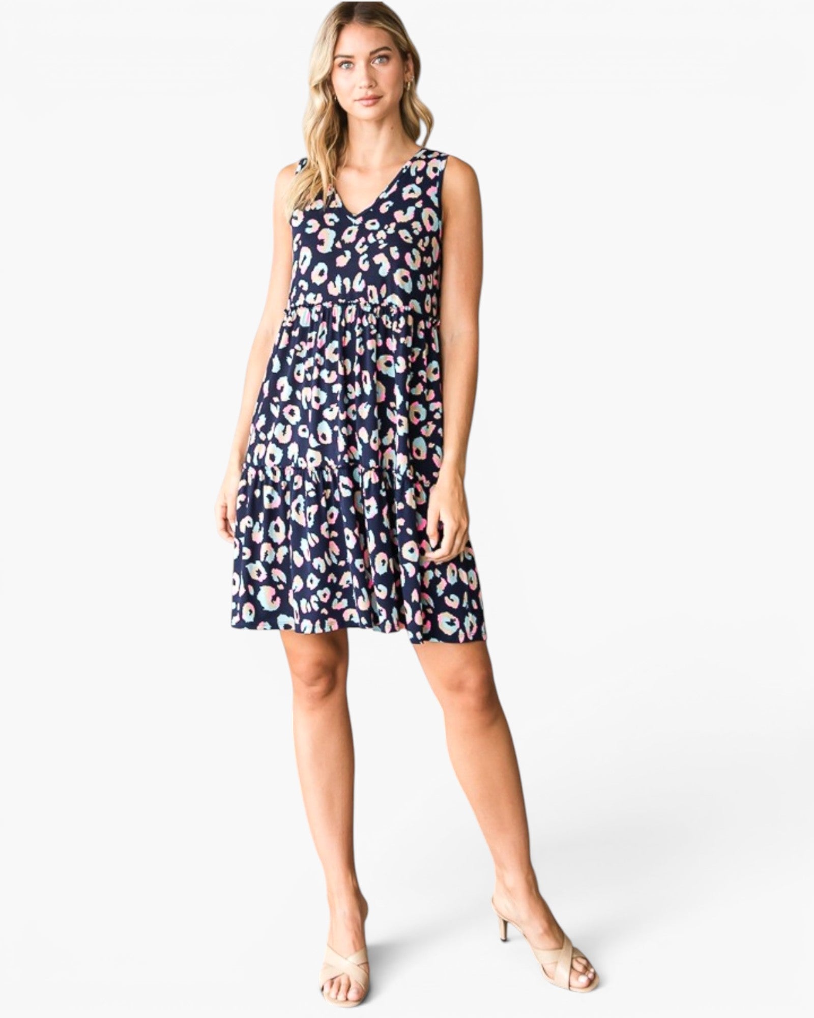 A woman modeling a sleeveless V-neck tiered dress with a navy blue base and a pink, white, and light blue abstract floral pattern, paired with clear high-heeled sandals against a light gray studio background.