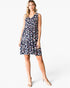 A woman modeling a sleeveless V-neck tiered dress with a navy blue base and a pink, white, and light blue abstract floral pattern, paired with clear high-heeled sandals against a light gray studio background.