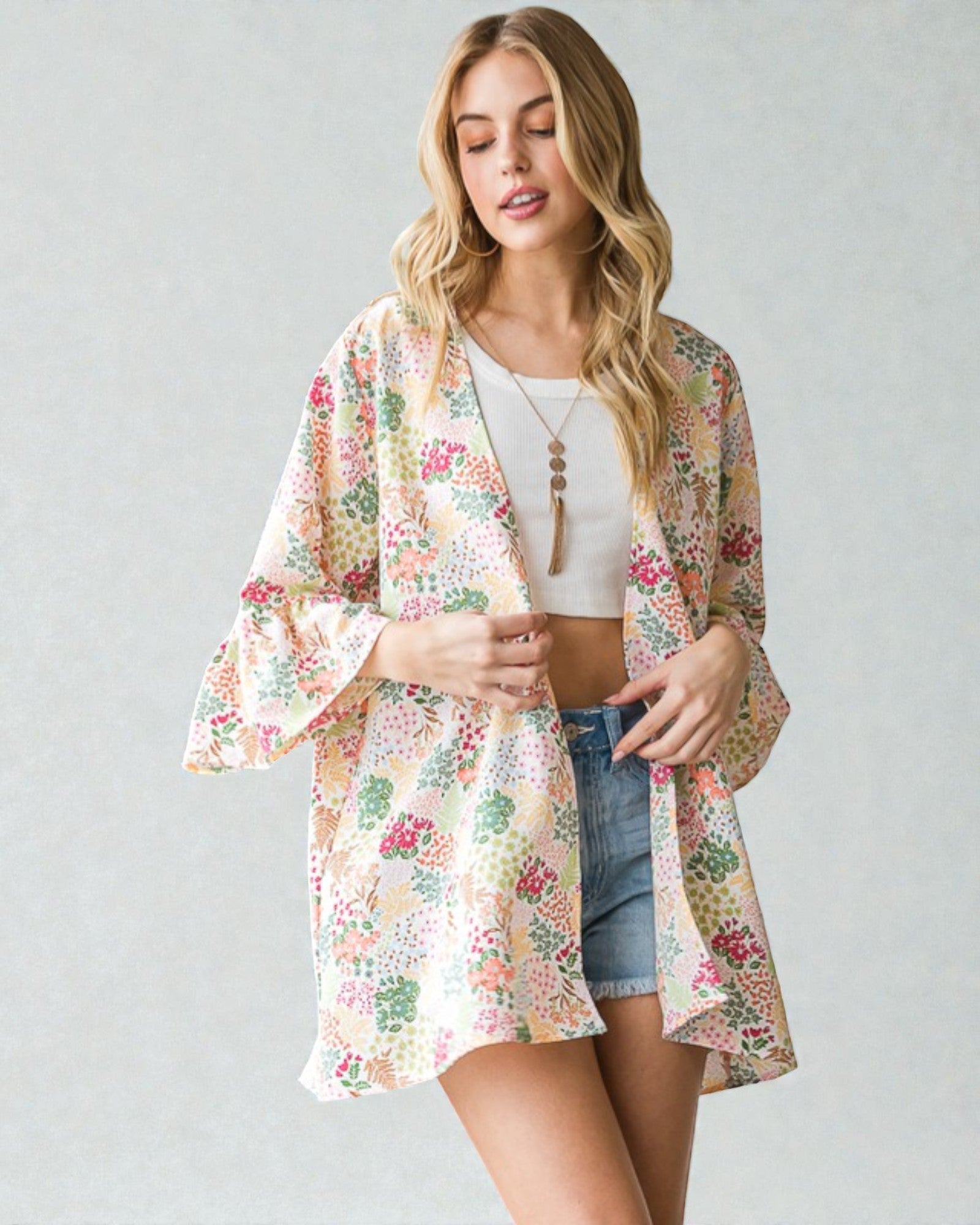Woman wearing a floral kimono over a white top and denim shorts on a plain background