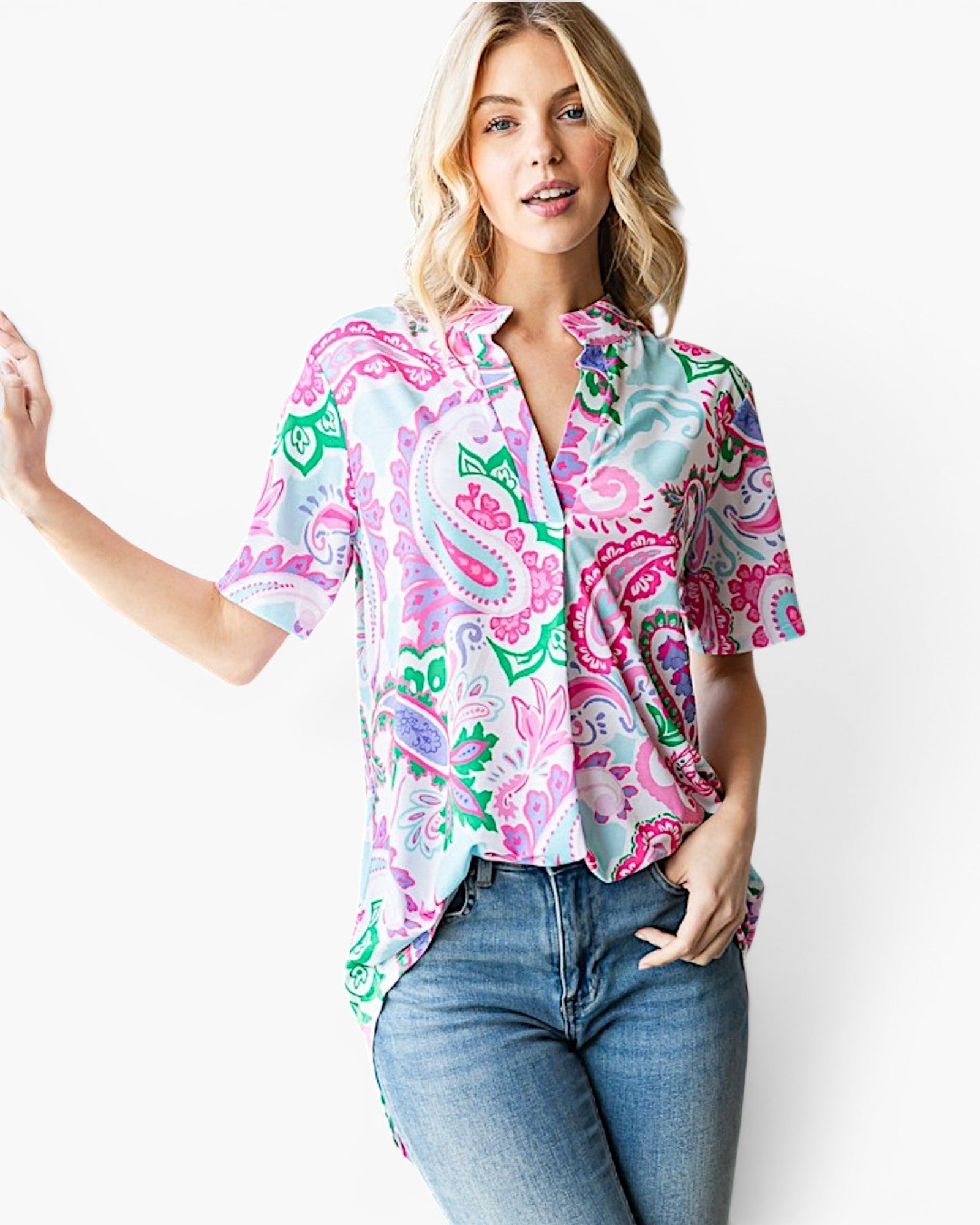 A woman modeling a short-sleeve button-up shirt with a vibrant paisley pattern in pink, green, and blue on a white base, paired with blue jeans against a plain white background.