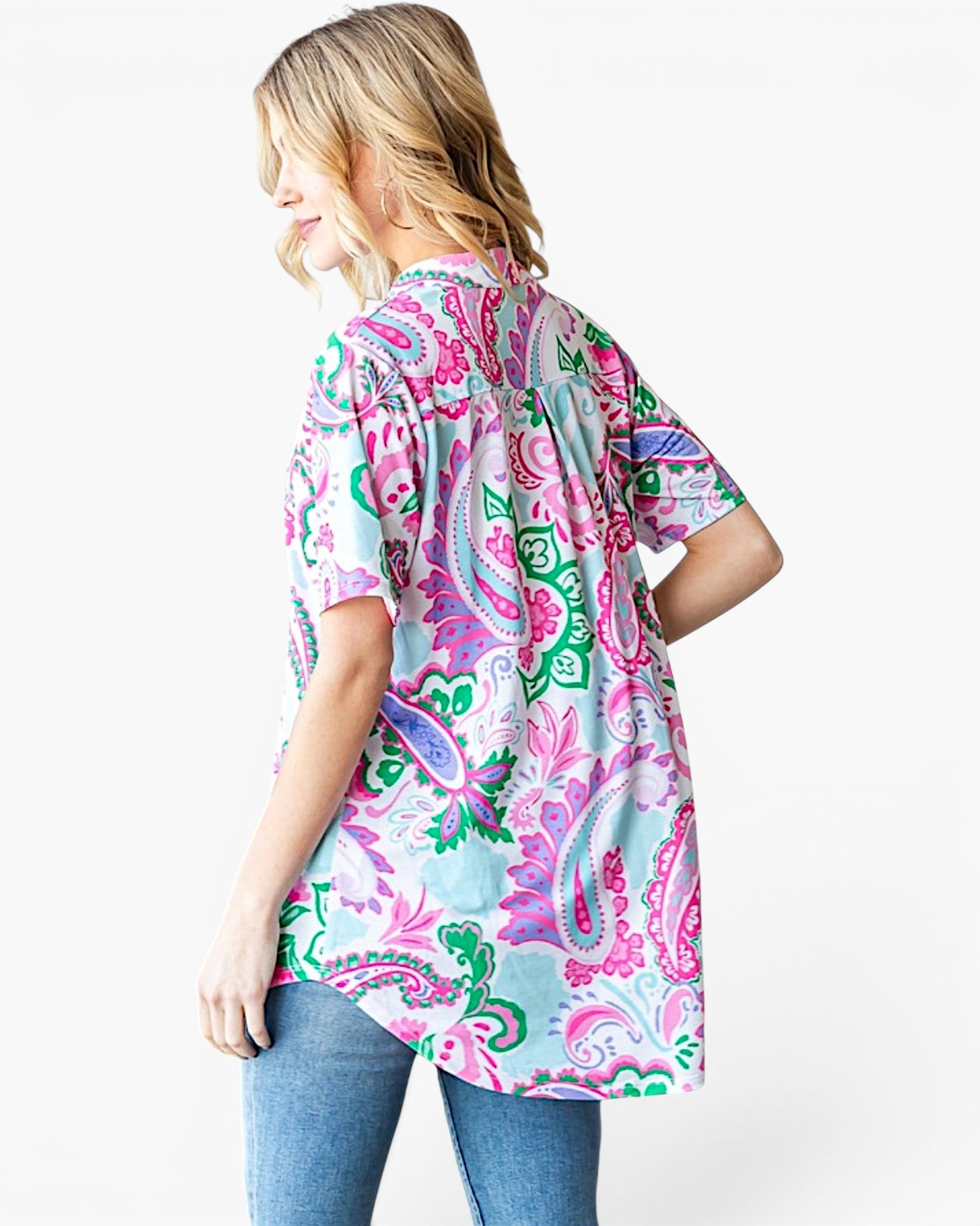 A woman wearing a short-sleeve button-up shirt with a colorful paisley pattern in pink, green, and light blue, paired with blue jeans, shown from a three-quarters back view against a plain white background.