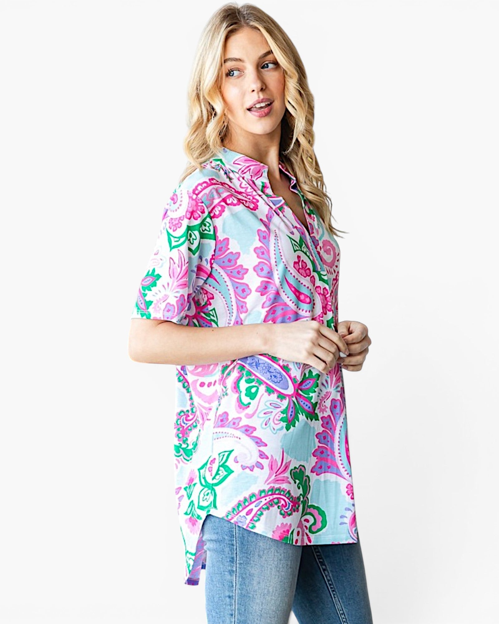 A woman modeling a short-sleeve button-up shirt with a vibrant paisley pattern in pink, green, and blue on a white background, paired with blue jeans against a plain white studio backdrop.