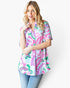 A woman modeling a short-sleeve button-up shirt with a colorful paisley pattern in pink, green, and blue on a white background, paired with blue jeans against a plain white studio backdrop.
