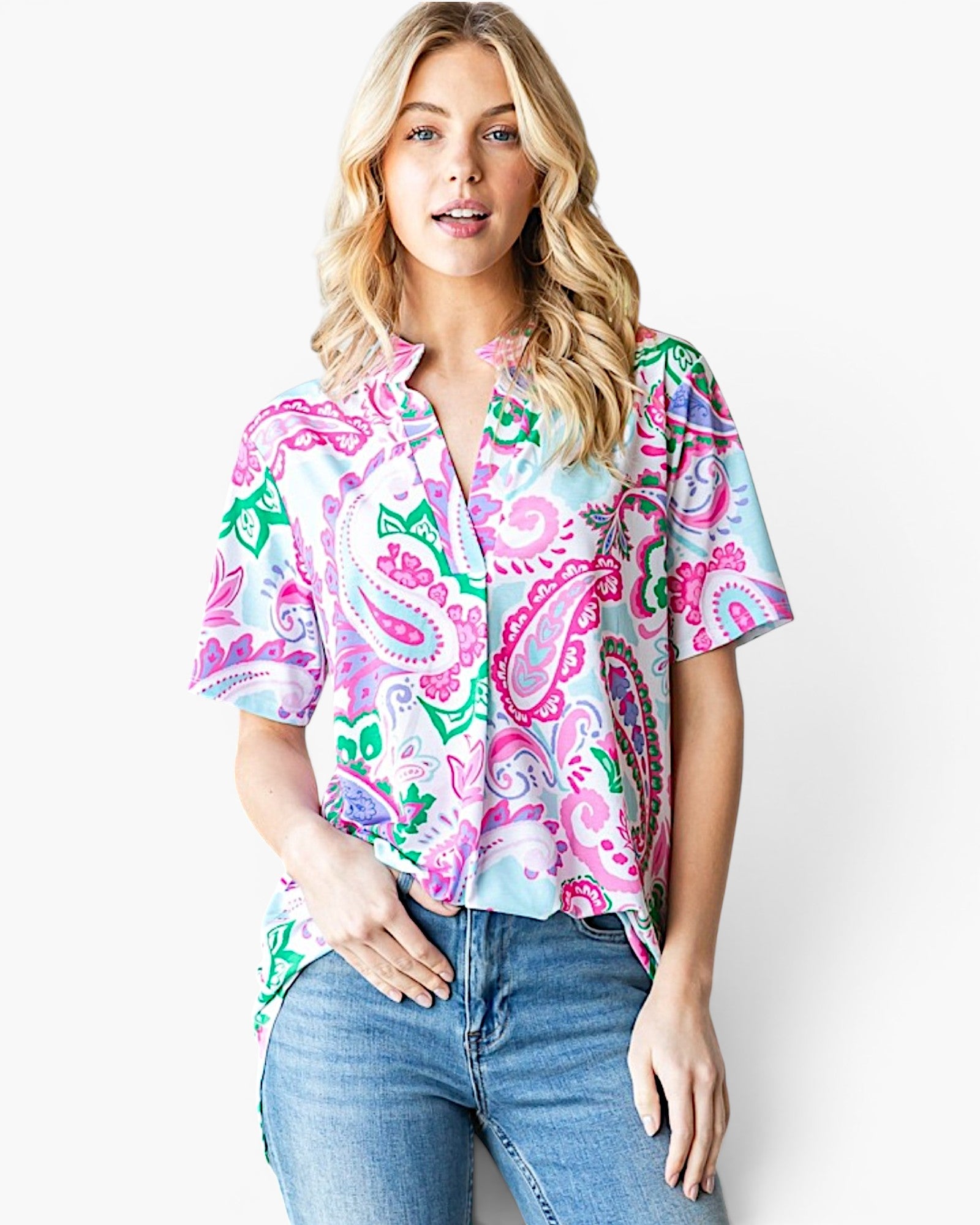 A woman modeling a short-sleeve button-up blouse with a vibrant paisley pattern in pink, green, and blue on a white base, paired with blue jeans against a plain white background.