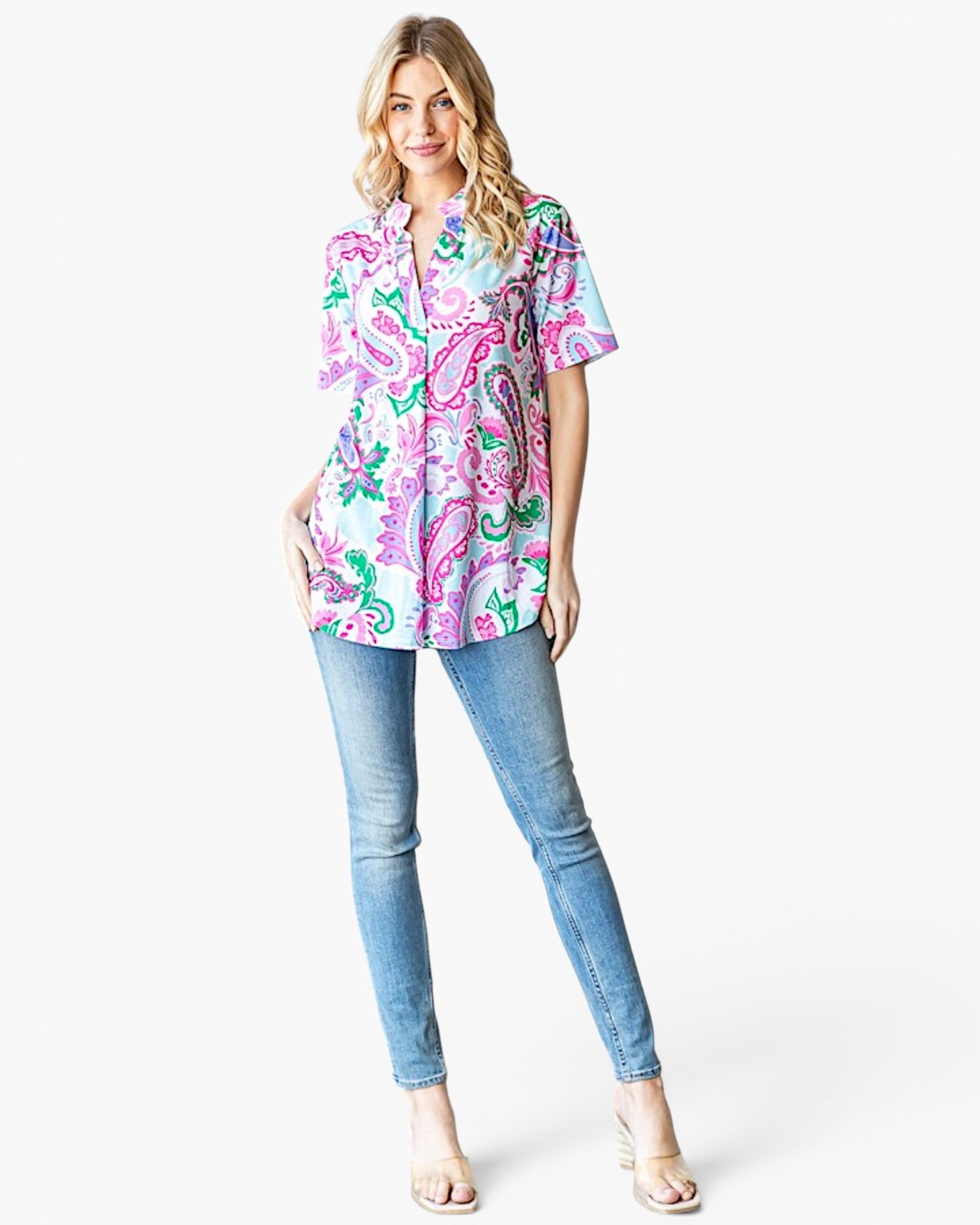 A woman modeling a short-sleeve button-up shirt with a colorful paisley pattern in pink, green, and blue on a white background, paired with light blue skinny jeans and beige sandals, against a plain white studio background.