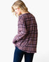 A woman wearing a pink and purple plaid long-sleeve top with dark blue jeans, photographed against a plain white background.