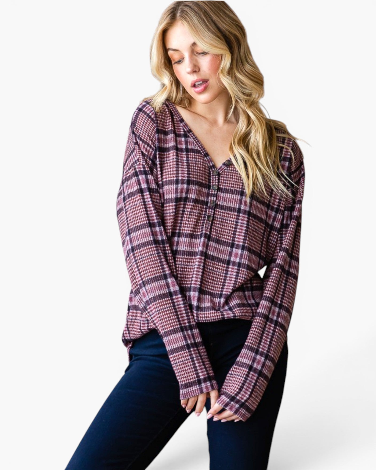 A woman modeling a long-sleeve plaid button-up shirt in a dusty pink, purple, and black pattern, paired with dark blue jeans, against a plain white studio background.