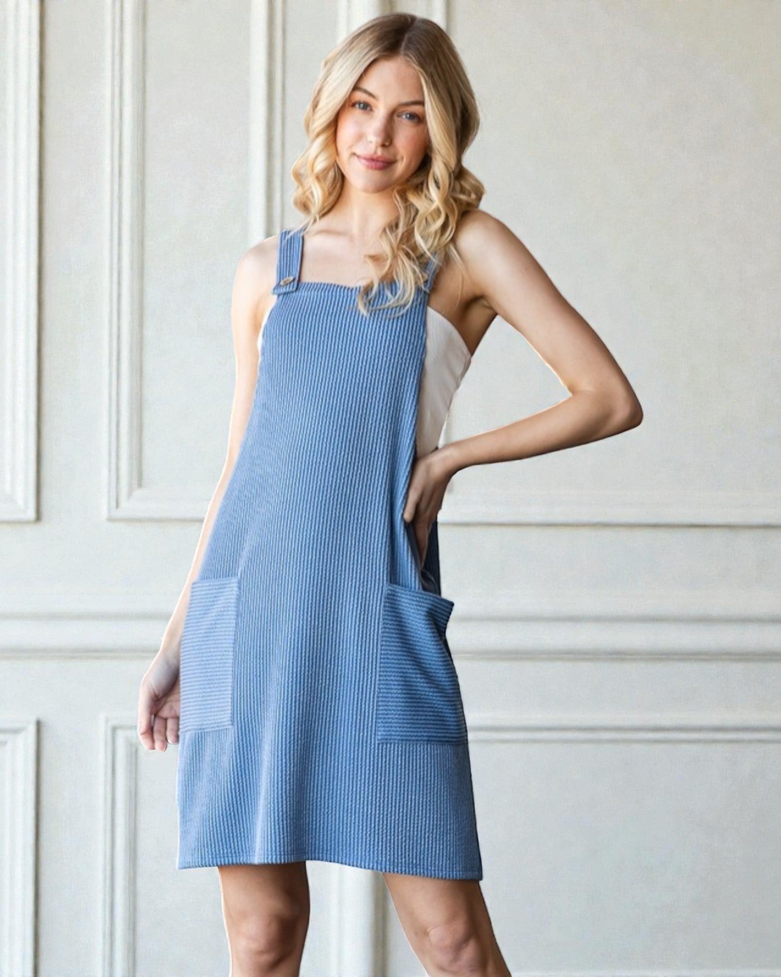 Woman wearing a blue pinafore dress against a white wall.