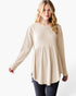 A woman modeling a beige long-sleeve ribbed knit top with a gathered peplum waist, paired with dark jeans, against a plain white studio background.