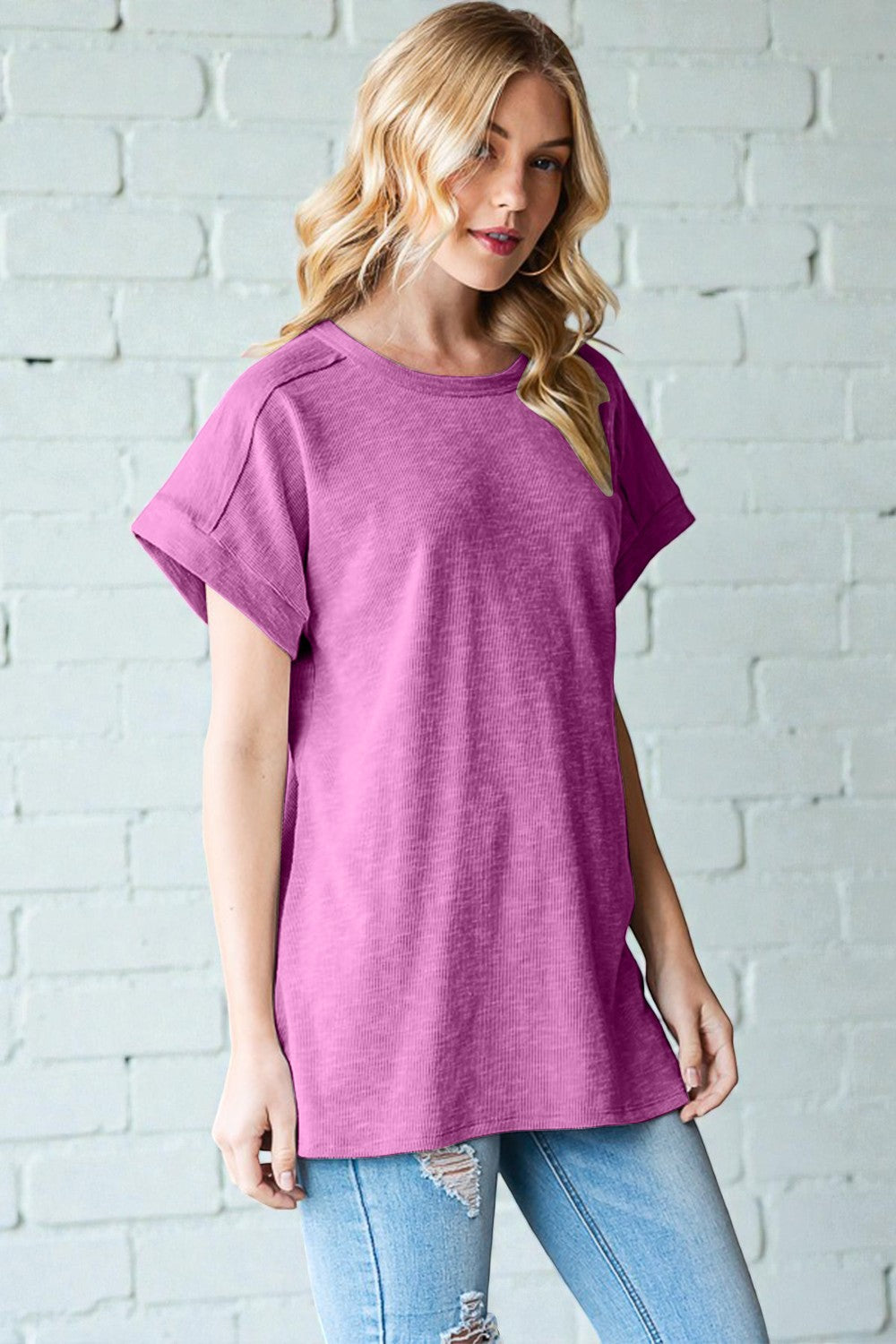 Woman wearing a purple t-shirt and light blue jeans against a white brick wall.