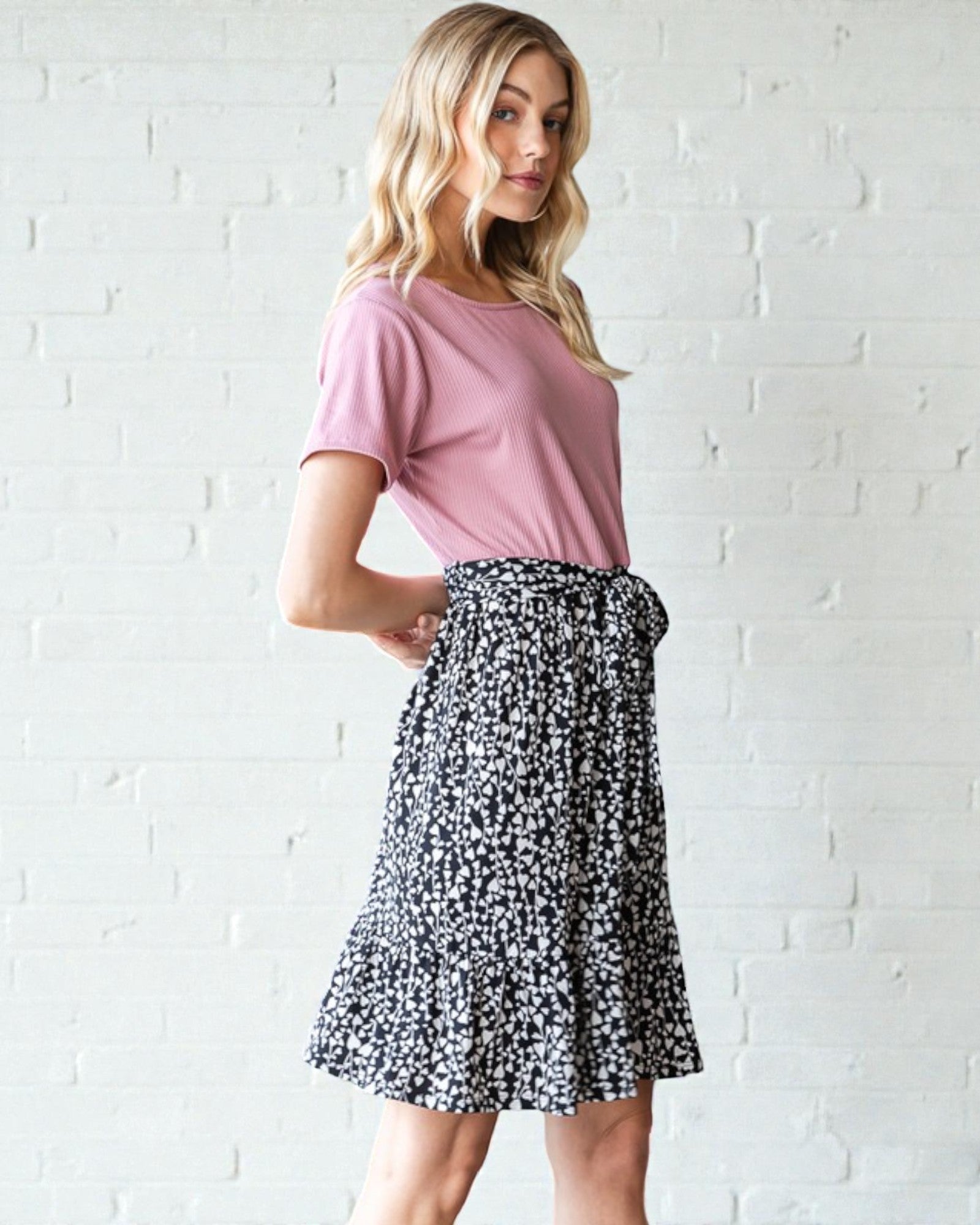 Woman wearing a pink top and black floral skirt against a white brick wall.