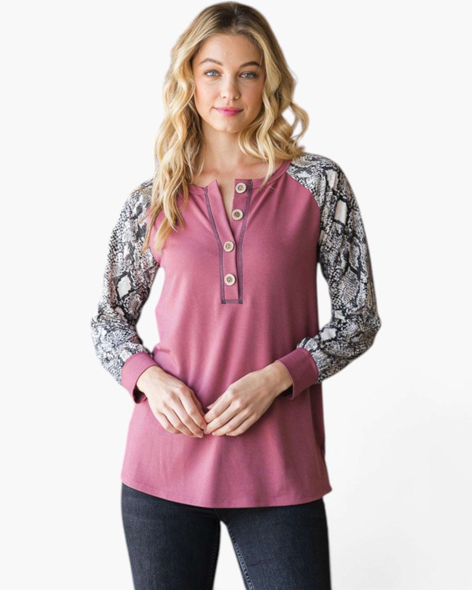 A woman modeling a dusty rose long-sleeve henley top with black and white snakeskin-patterned sleeves, paired with dark jeans, against a plain white background.