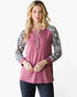 A woman modeling a dusty rose long-sleeve henley top with black and white snakeskin-patterned sleeves, paired with dark jeans, against a plain white background.