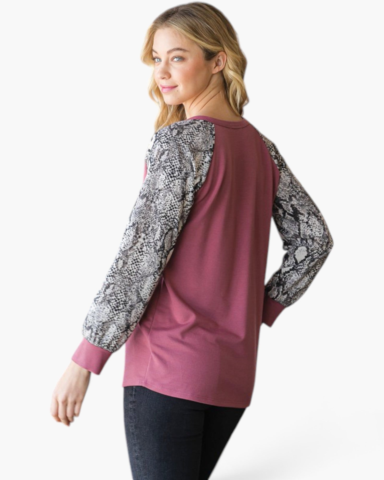 A woman wearing a dusty rose long-sleeve top with black and white snakeskin-patterned sleeves, shown from a three-quarter back angle against a plain white studio background.