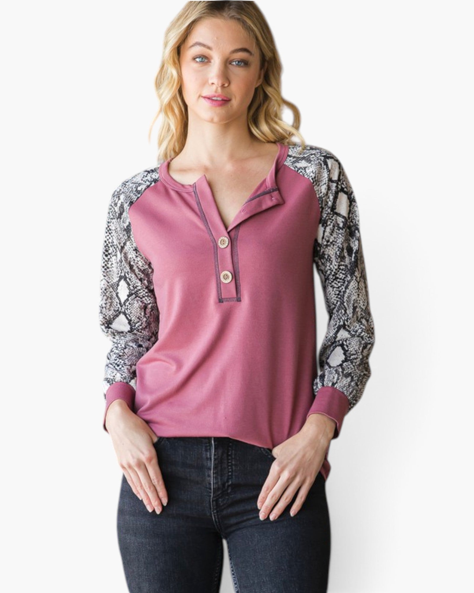 A woman modeling a long-sleeve henley top with a dusty rose body and black-and-white snakeskin patterned sleeves, paired with dark jeans against a plain white background.