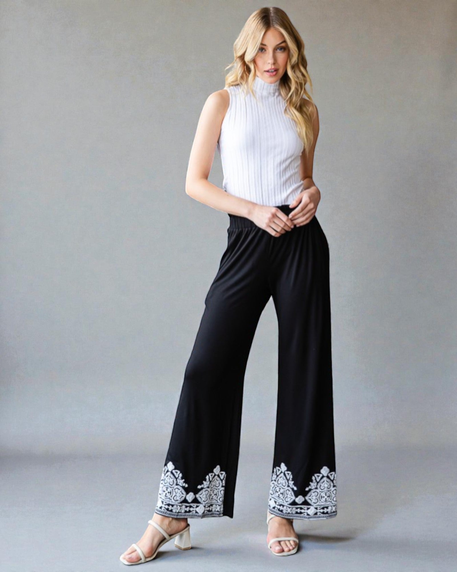 Woman wearing a white sleeveless top and black pants with lace details on a gray background