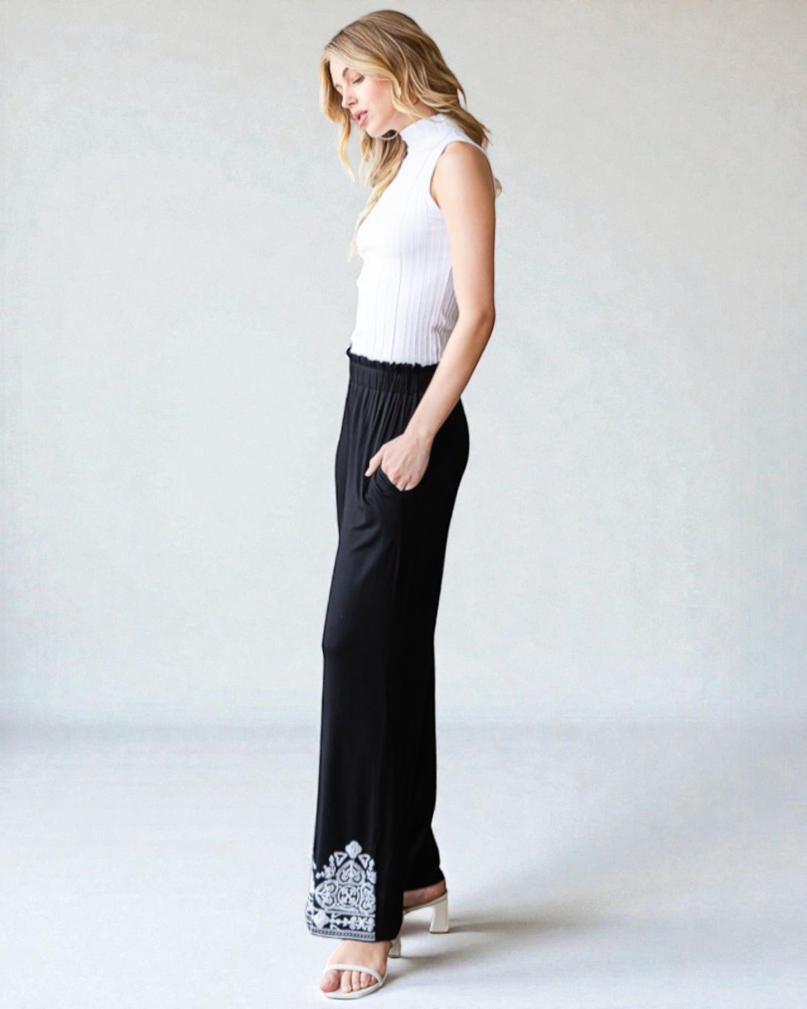 Woman wearing a white sleeveless top and black pants with floral embroidery on a plain background