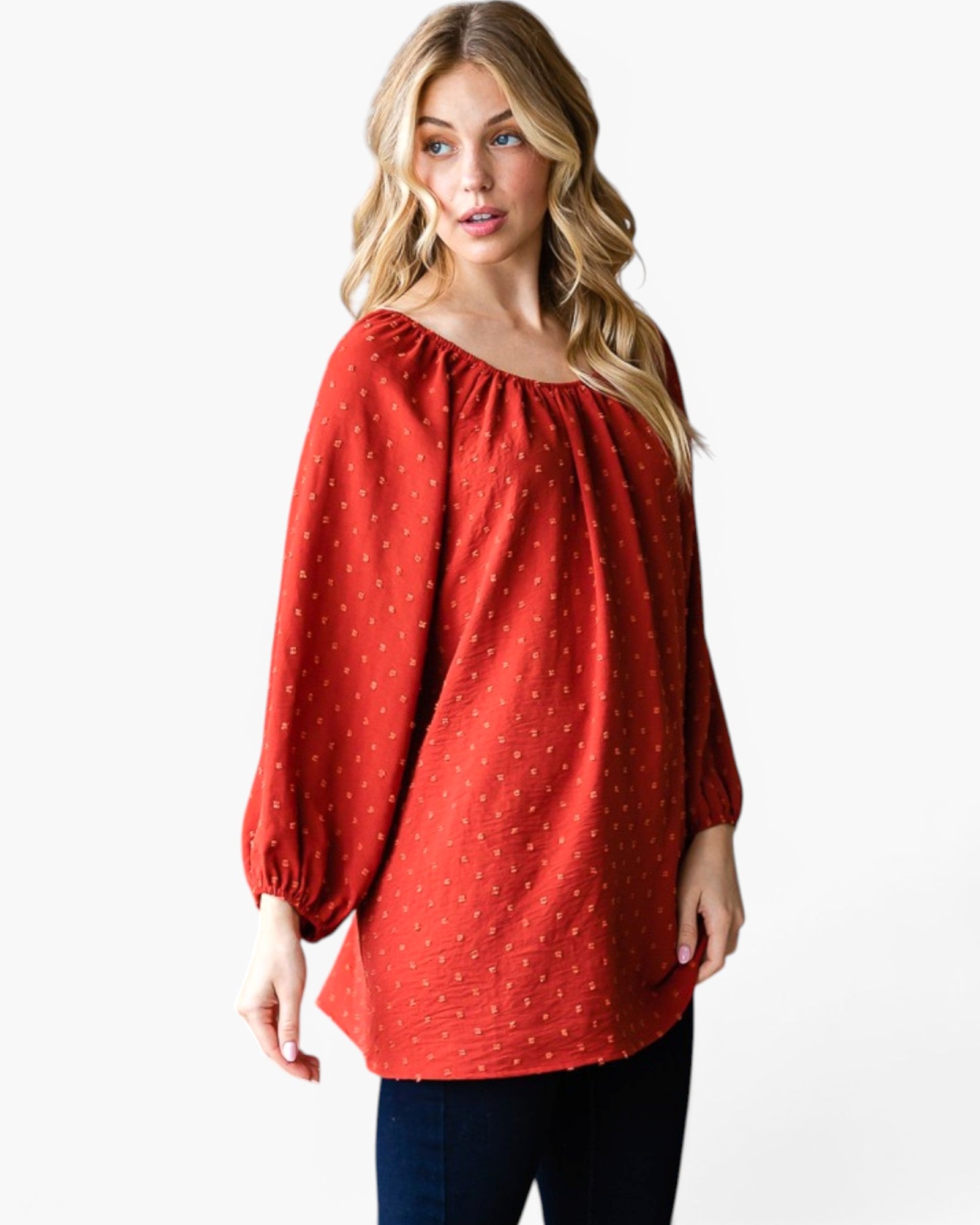 A woman wearing a rust red long-sleeve blouse with a small white polka dot pattern, paired with dark blue jeans, photographed against a solid white studio background.
