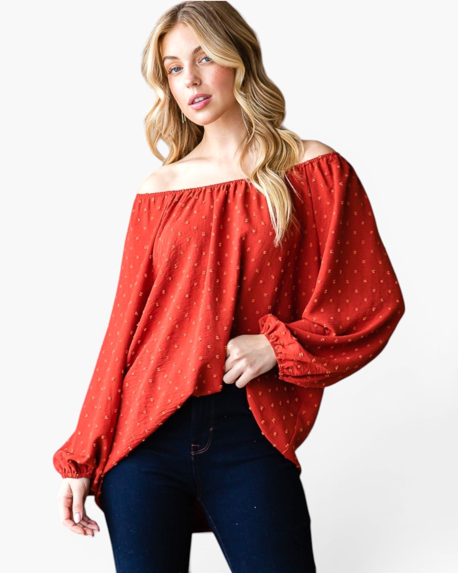 A woman wearing a rust red off-the-shoulder blouse with a tiny white polka dot pattern and dark blue jeans, photographed against a clean white studio background.