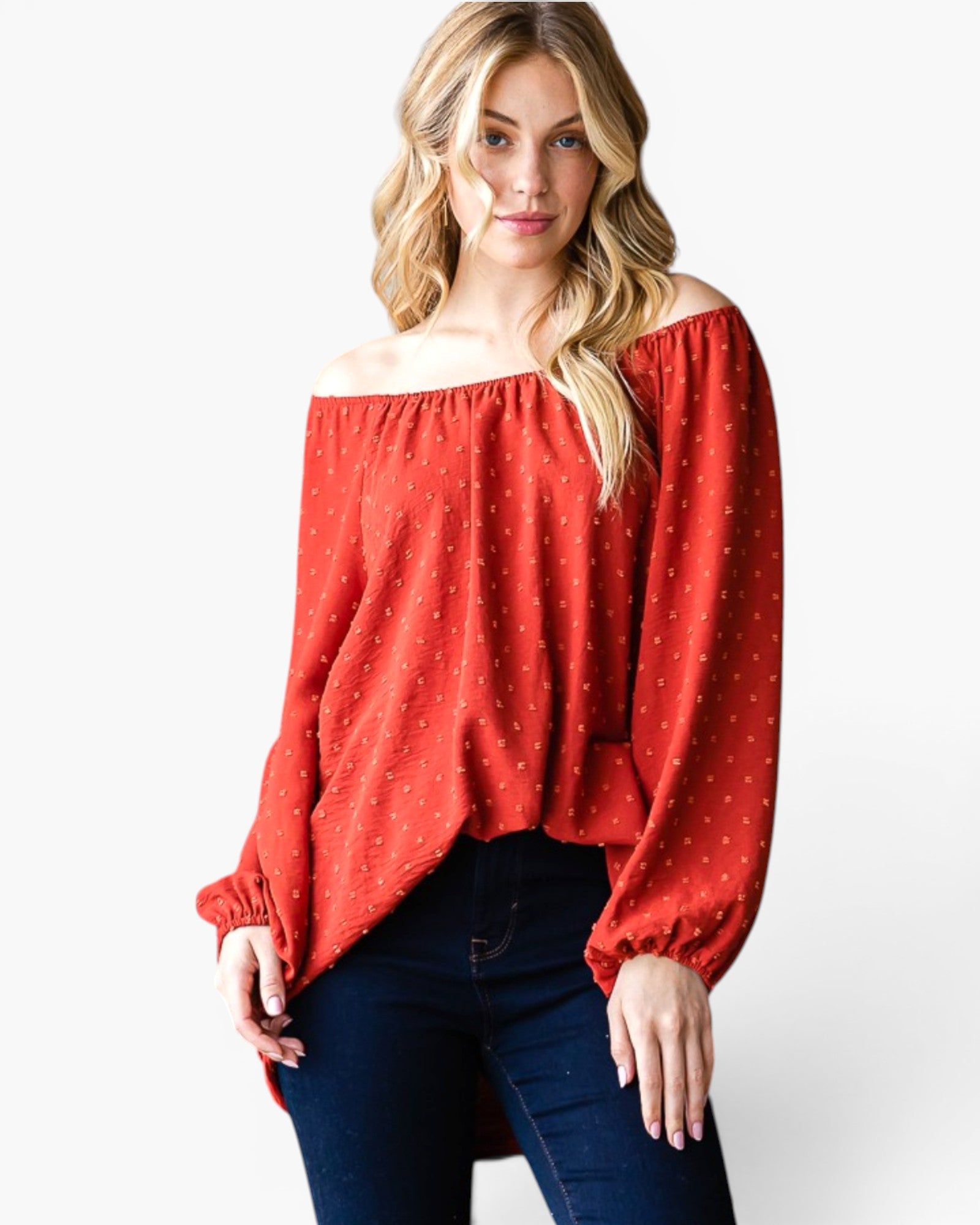 A woman wearing a rust red off-the-shoulder long-sleeve blouse with a tiny white polka dot pattern, paired with dark blue jeans, against a plain white studio background.