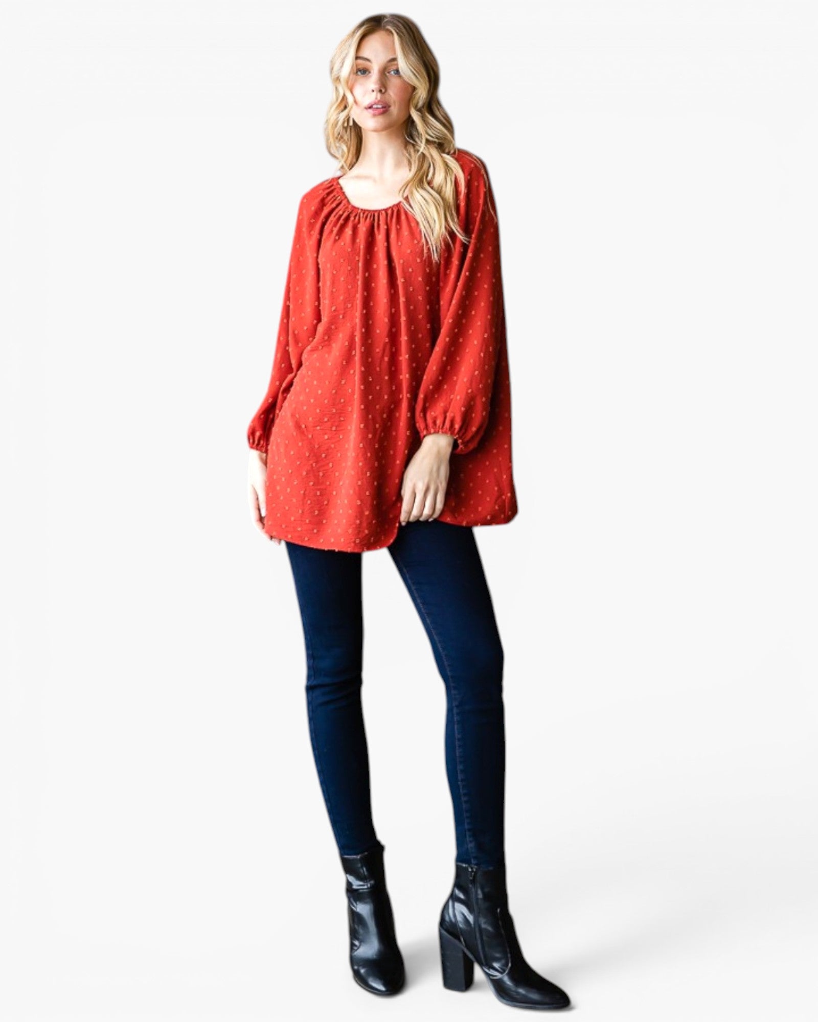 A woman modeling a textured red long-sleeve tunic top with a gathered neckline, paired with dark blue skinny jeans and black ankle boots, against a plain white studio background.