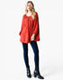 A woman modeling a textured red long-sleeve tunic top with a gathered neckline, paired with dark blue skinny jeans and black ankle boots, against a plain white studio background.