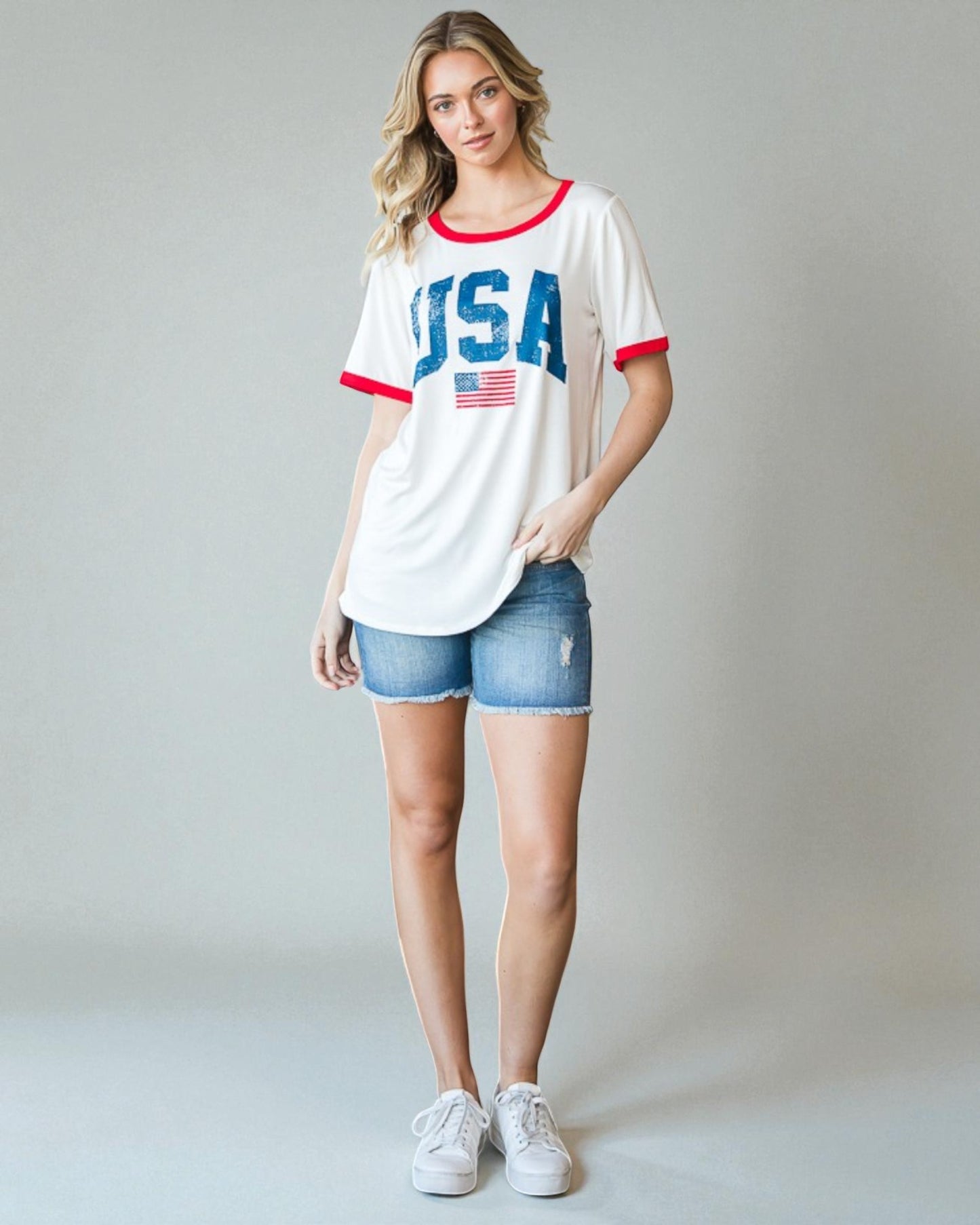 Woman wearing a white 'USA' t-shirt with red and blue accents, denim shorts, and white sneakers on a gray background.