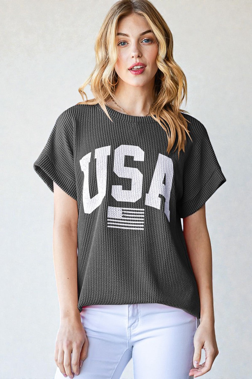 Woman wearing a dark gray 'USA' graphic t-shirt with white pants on a light background