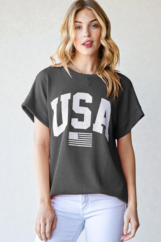 Woman wearing a dark gray 'USA' graphic t-shirt with white pants on a light background